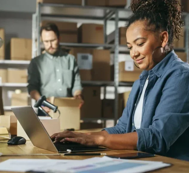 Lady-types-on-a-keyboard-in-her-warehouse-while-a-colleague-moves-boxes